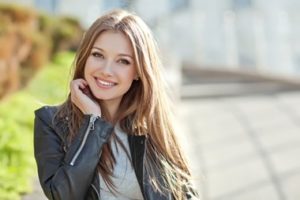 Young woman wearing a leather jacket and a grey top with her hand under her chin is smiling