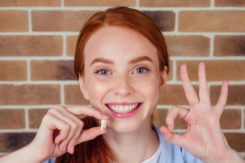 Woman with red hair making "ok" sign with one hand and holding extracted tooth with the other